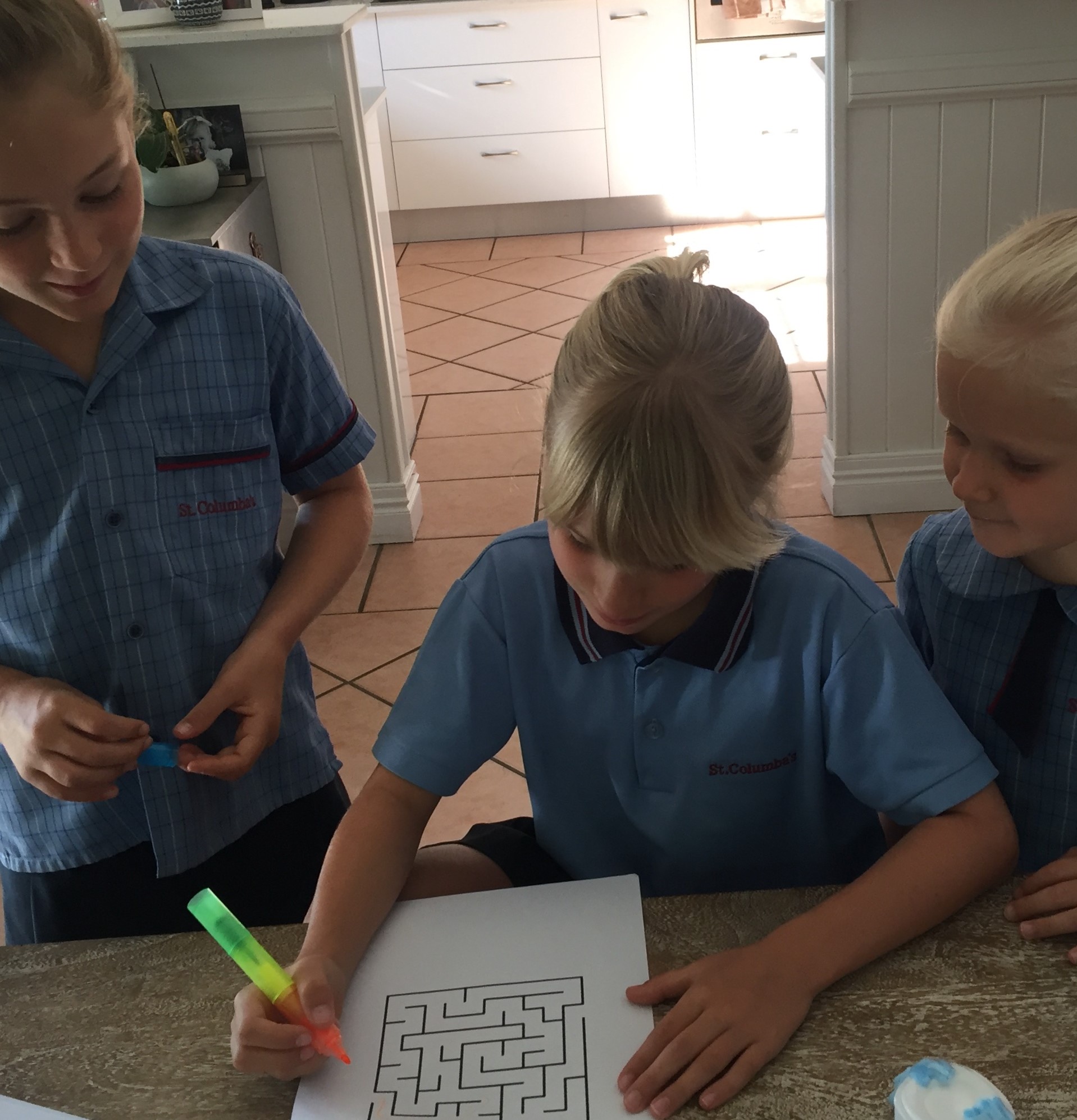 Three primary school students in blue uniforms working together at a table while one student completes a maze activity on paper with a highlighter