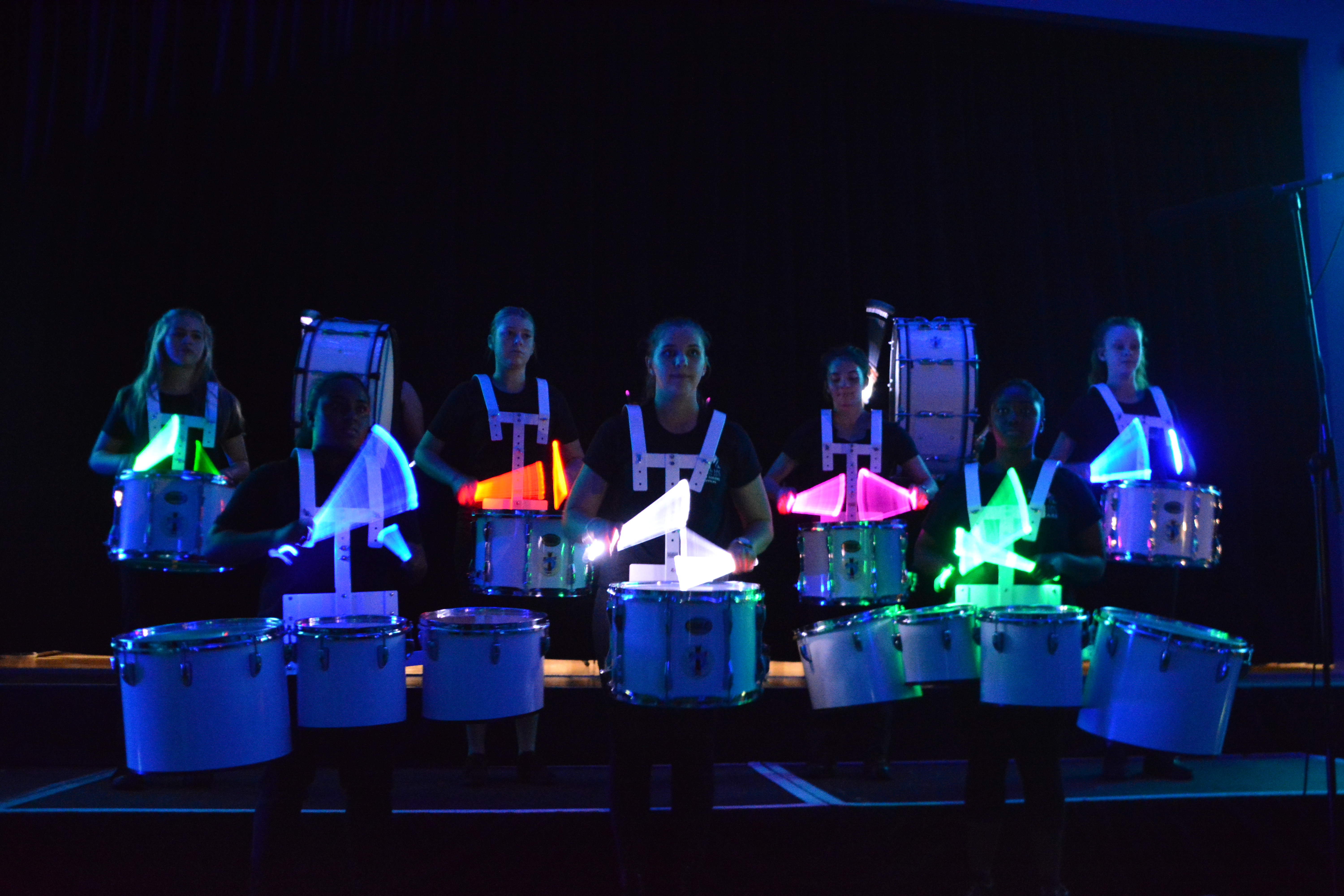 Students performing in a drumline on stage, wearing illuminated harnesses and playing drums under blue stage lighting