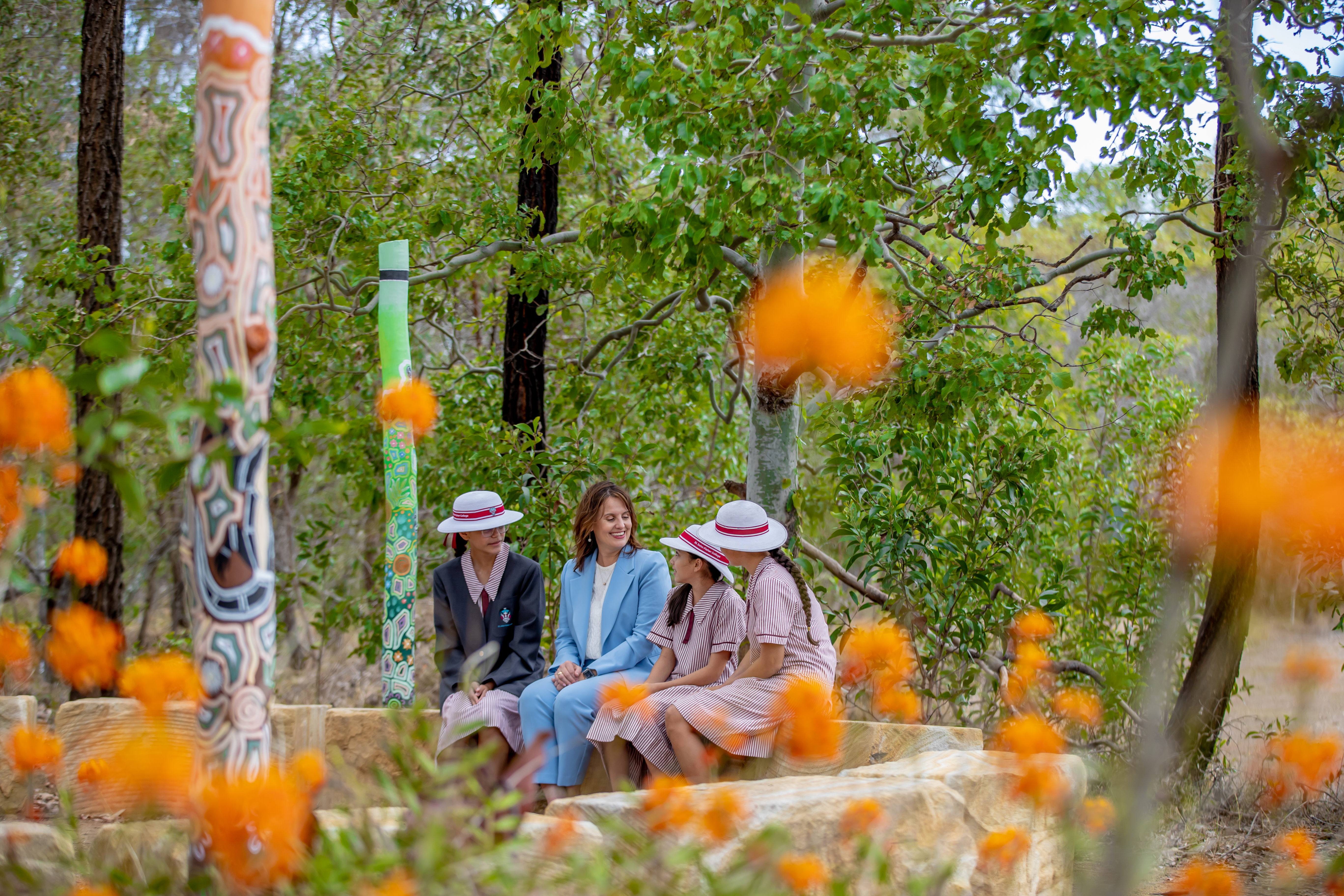 Students and an adult sitting together on stone seating in a garden area with decorated poles and surrounding trees