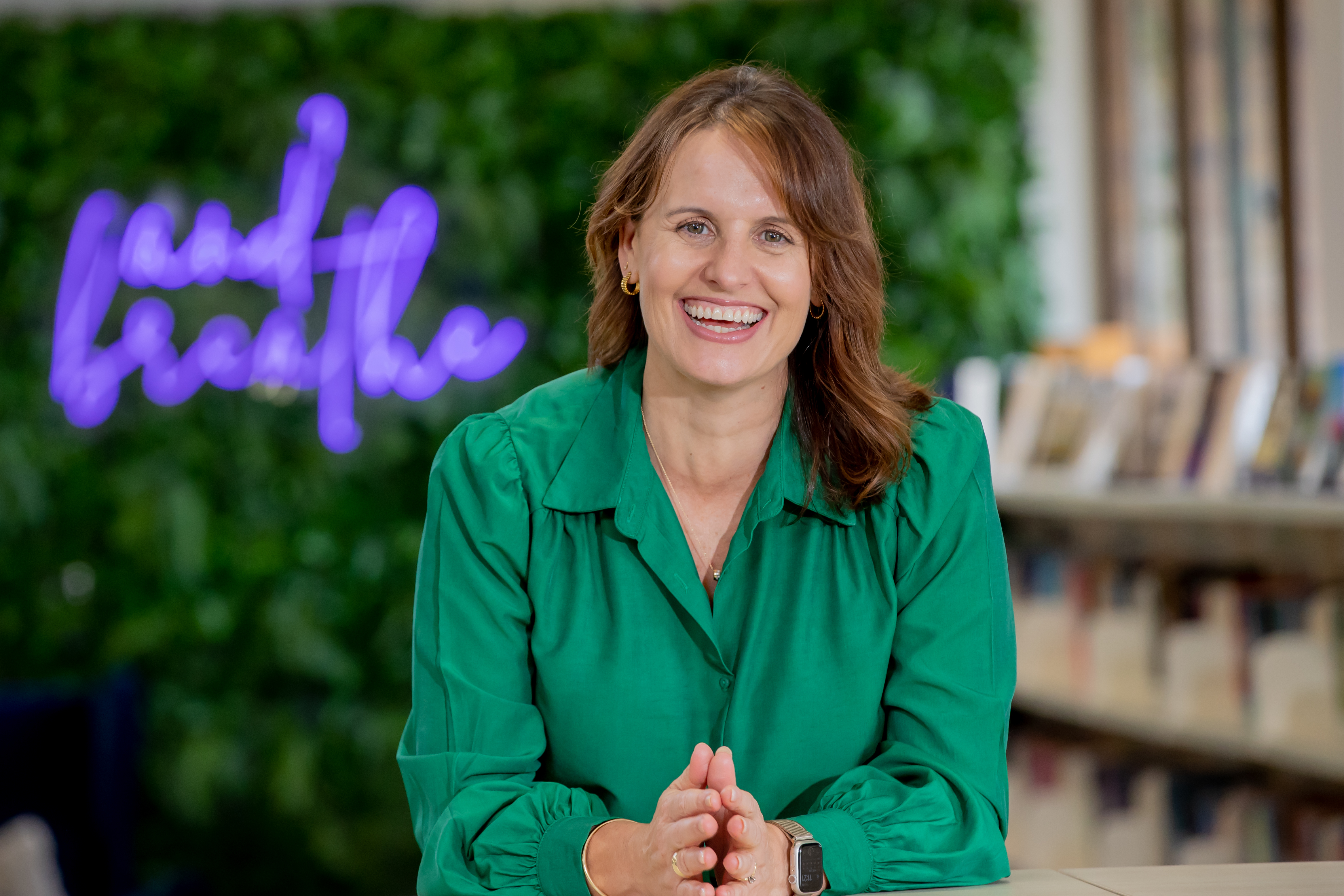 School principal Britt Gurnett smiling while seated indoors with bookshelves and a green wall in the background