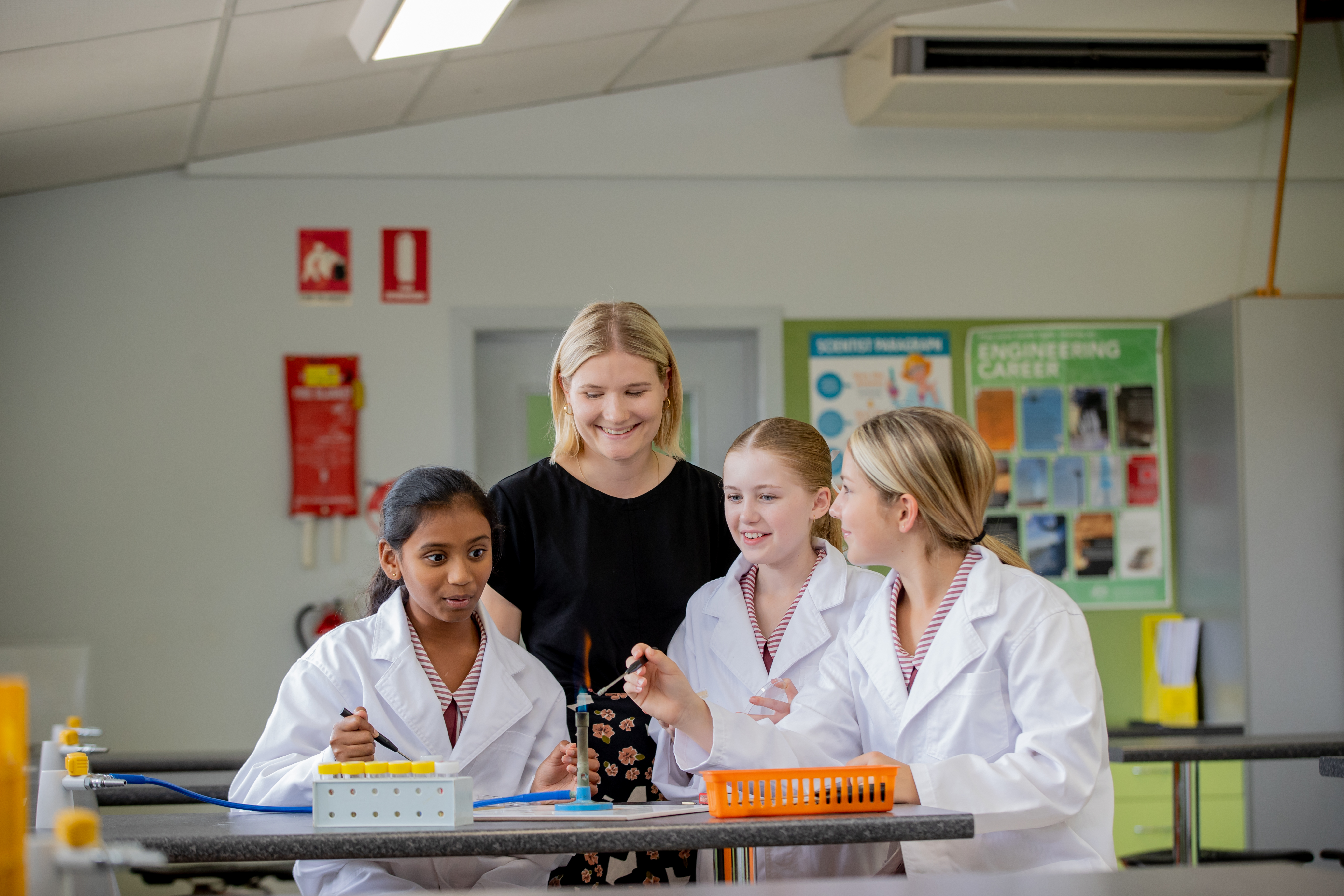 Four students wearing lab coats work together at a science bench in a classroom, using equipment and discussing an experiment while posters and safety signs are visible in the background