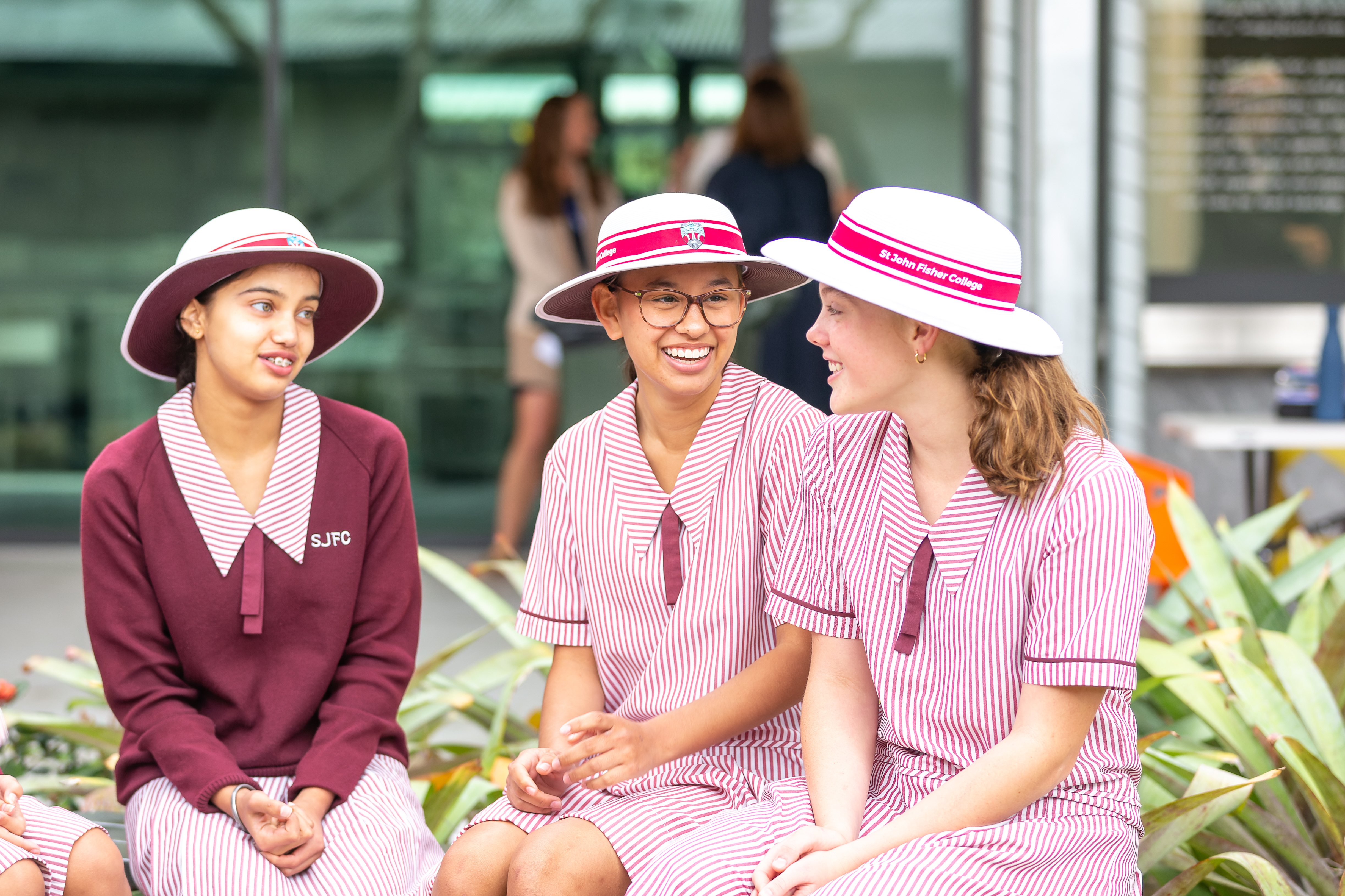 Three students in pink-and-white striped school uniforms and wide-brimmed hats sit together outdoors on a low wall with school buildings and plants in the background