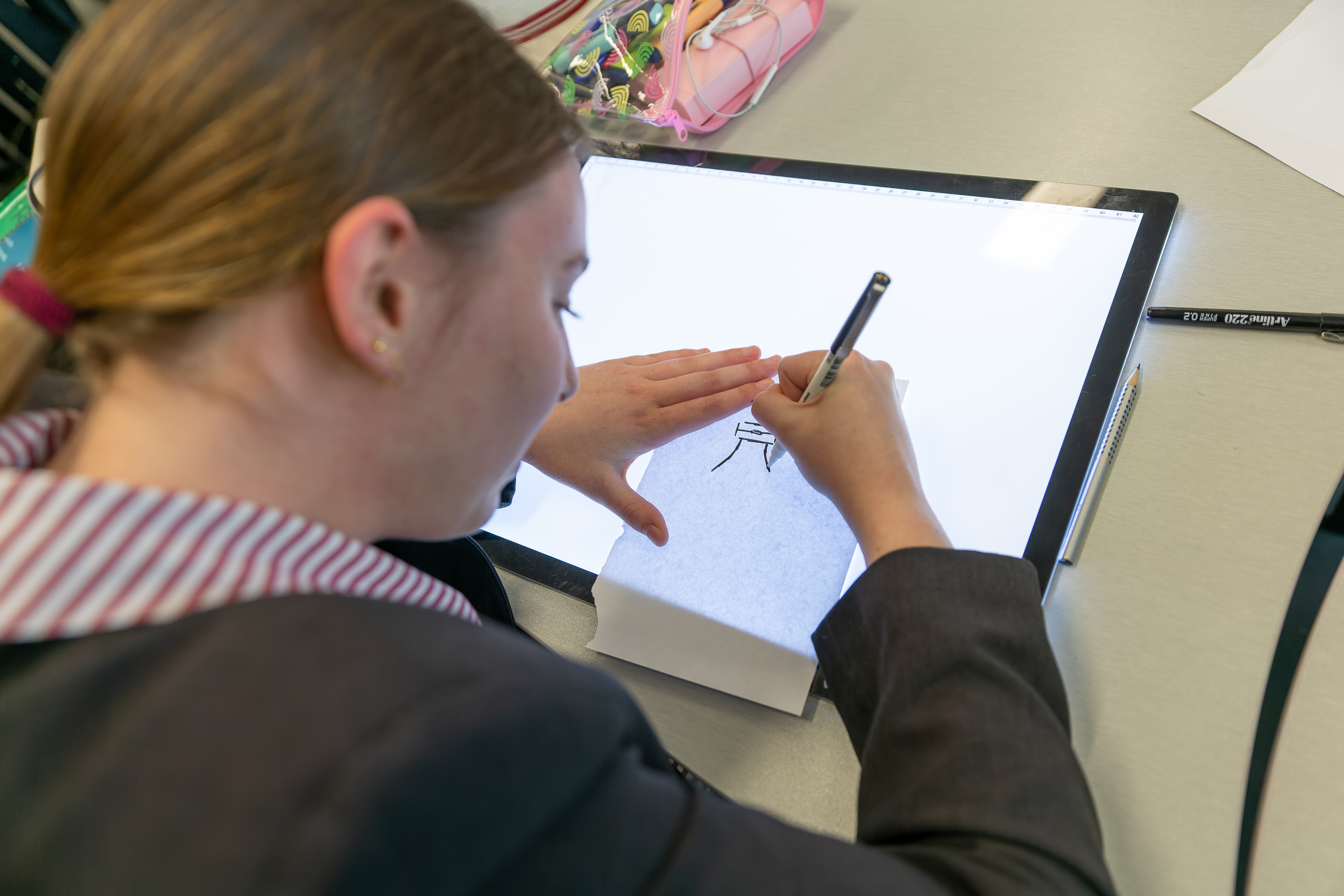 Student in school uniform using a lightbox to trace and draw an illustration at a classroom desk